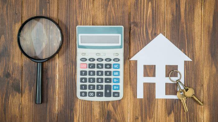 A magnifying glass, calculator, cutout of a house and some keys on a dark wood surface