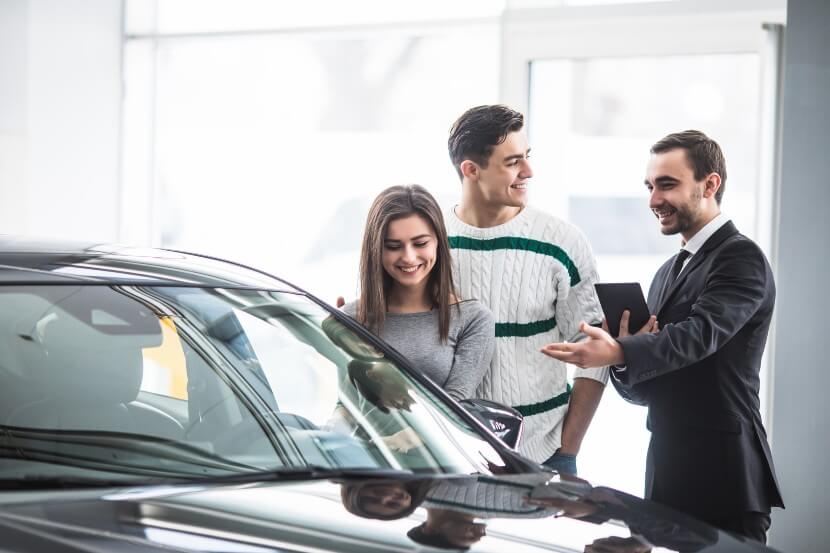 Young couple choosing a car at the dealership talking to a car salesman with tablet in hands