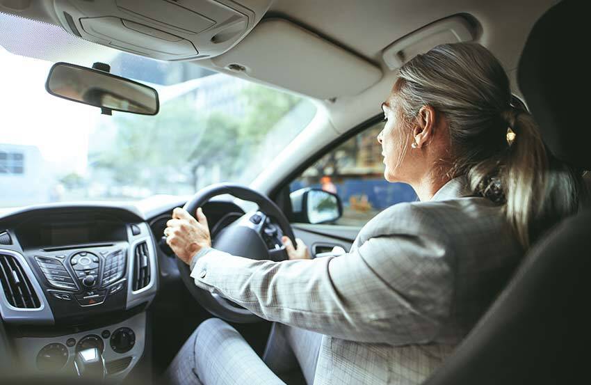 Shot taken from the back seat of a woman in a light grey suit driving a car