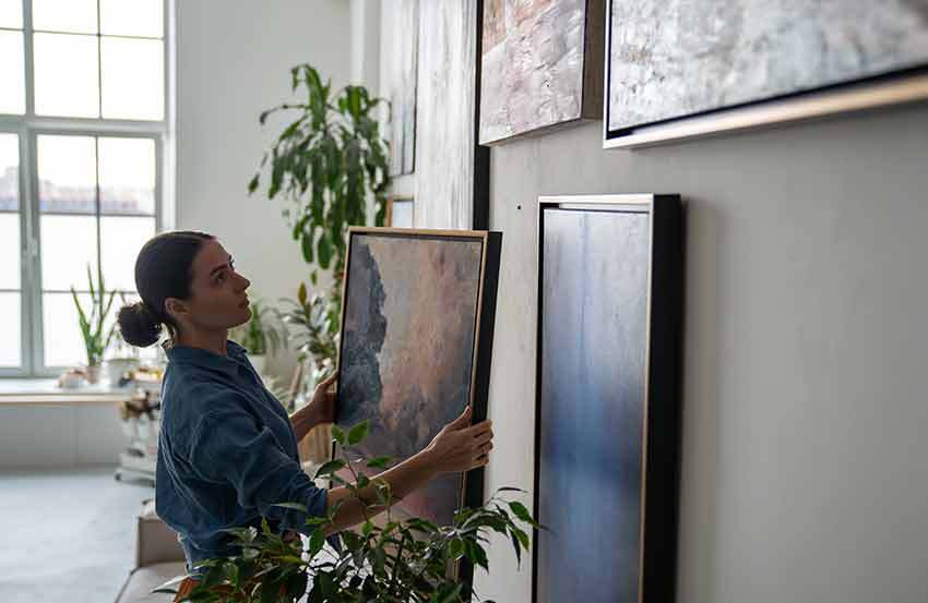 Woman hanging pieces of art on a wall, with houseplants in the background