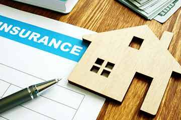 Wooden cutout of a house holding down an insurance document on a wooden table with a pen visible