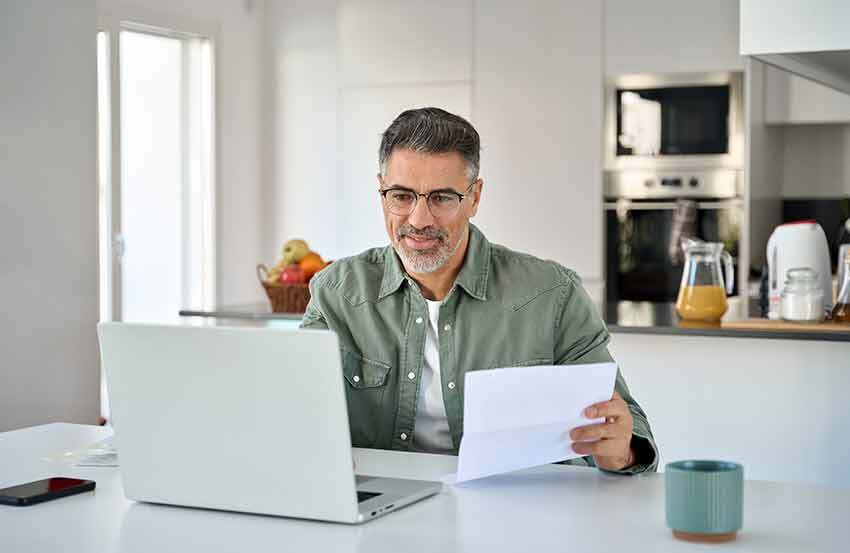 A man sat looking at a laptop holding a piece of paper