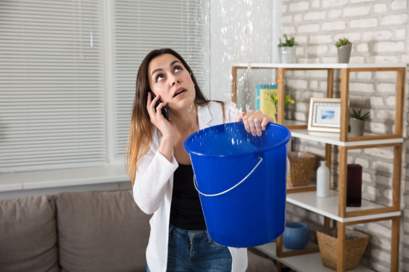 Woman standing in living room with a bucket, catching water from a burst pipe and on the phone to an emergency plumber