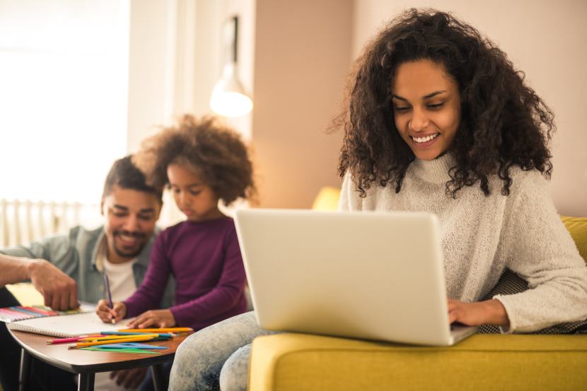 woman smiling working on her laptop while partner plays with their child in the background