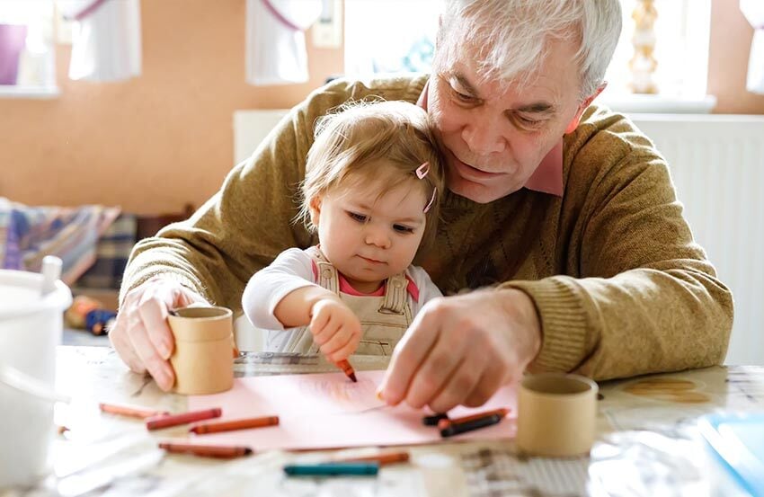 An older man and a young girl happily coloring together at a table, surrounded by crayons and colorful drawings.