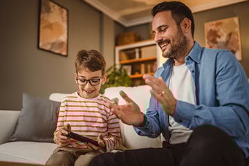 A father and son joyfully playing video games together, sharing smiles and excitement in their living room.
