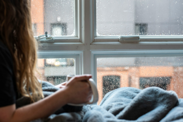 Female with blanket holding mug looking out of rainy window