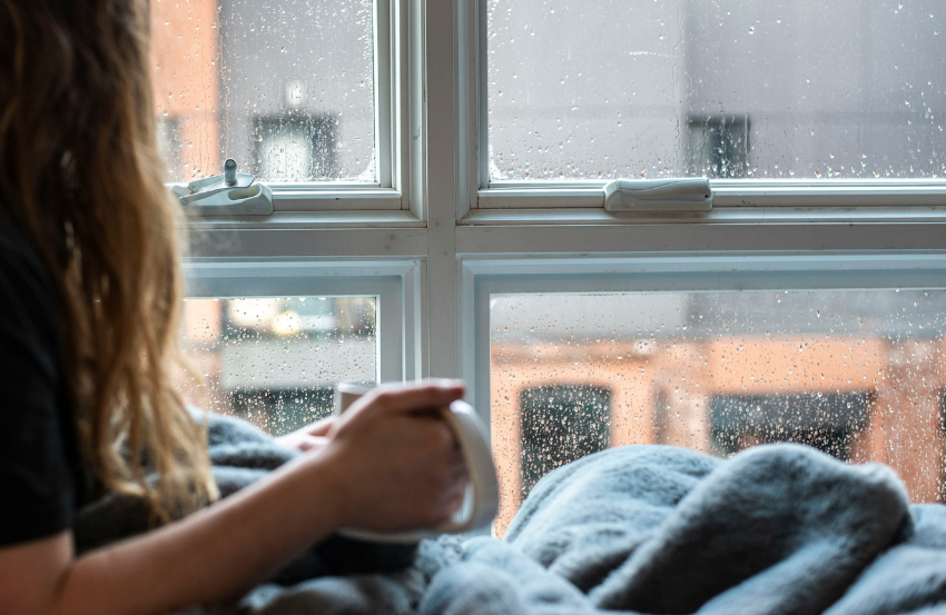 Female with blanket holding mug looking out of rainy window
