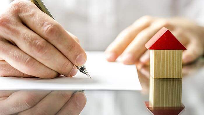 Hand signing agreement with wooden model house in foreground