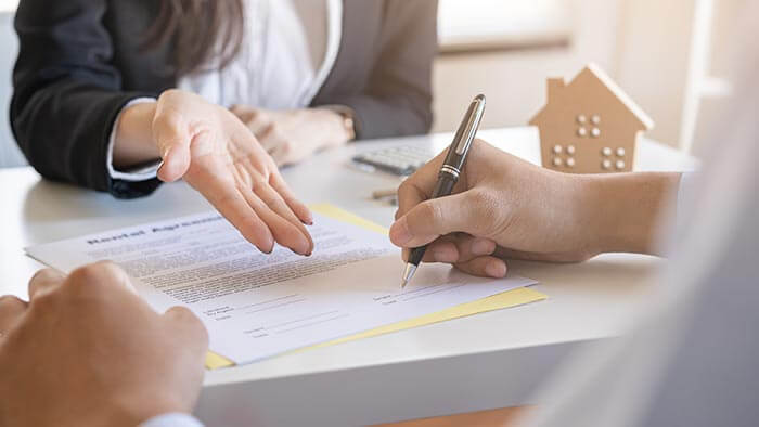 Close up of a hand signing a mortgage agreement while a lender gestures at the paper.