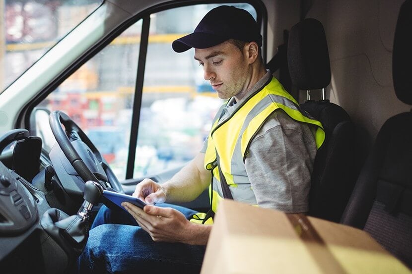 A delivery driver sitting in a van alongside a parcel checking paperwork on a tablet computer